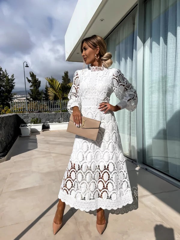 Woman in a white lace dress standing on a rooftop with modern architecture and greenery in the background.