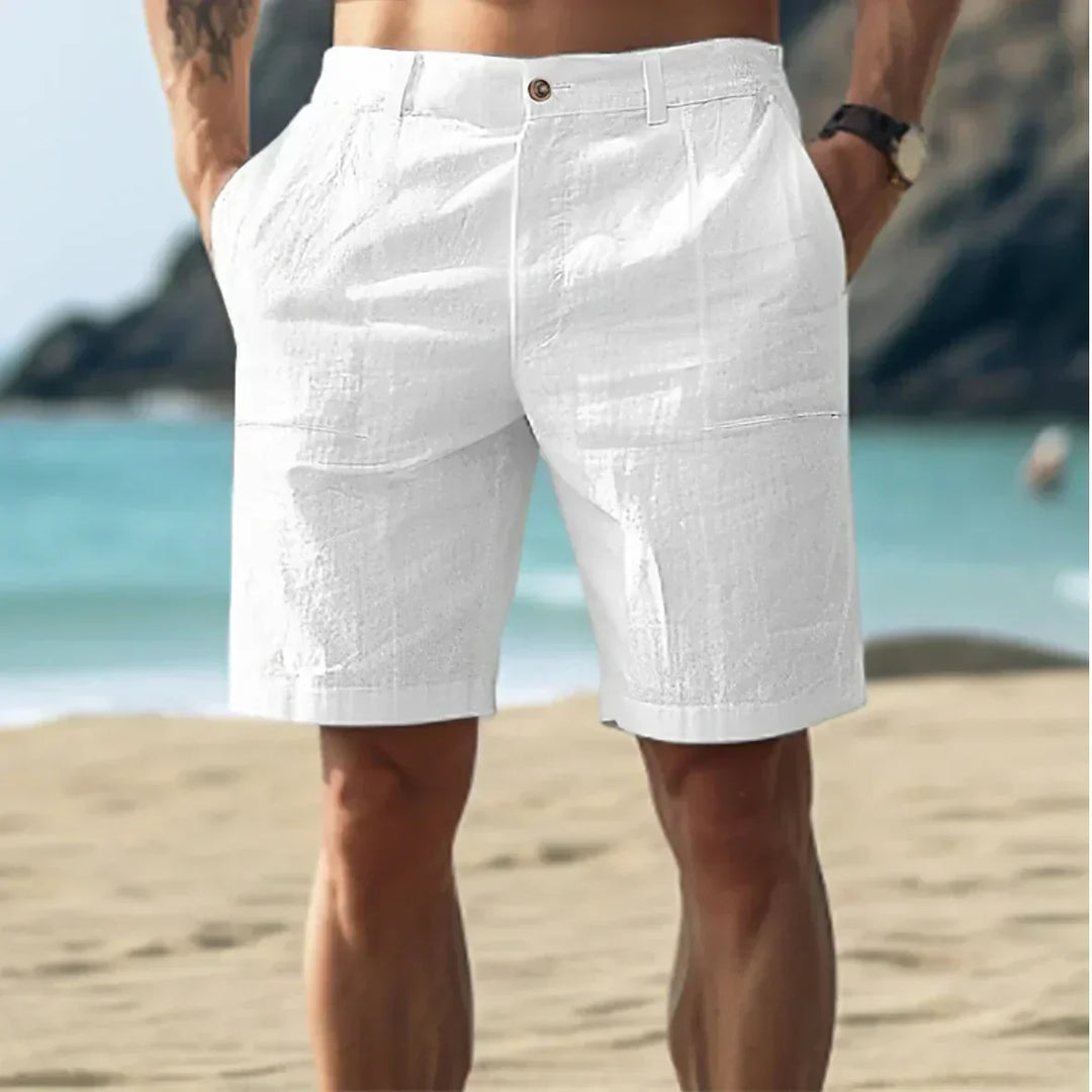 Man wearing white men's relaxed chino shorts with button closure on beach, rear view showing straight-cut silhouette and side pockets