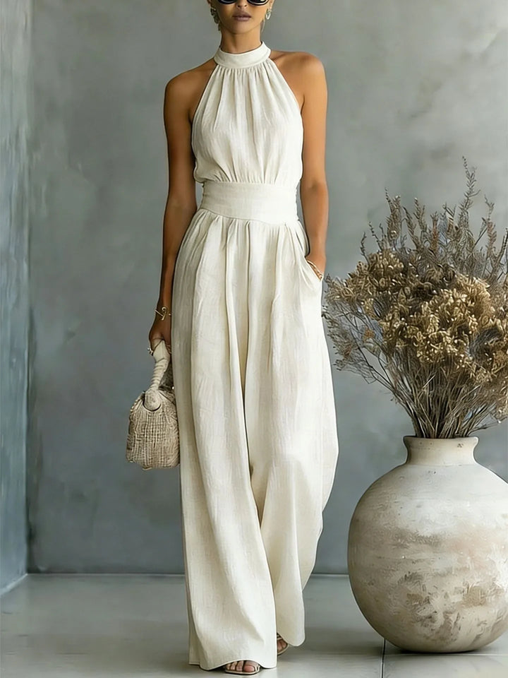 Halter wide leg jumpsuit in cream with cinched waist and flowing silhouette worn with woven bag